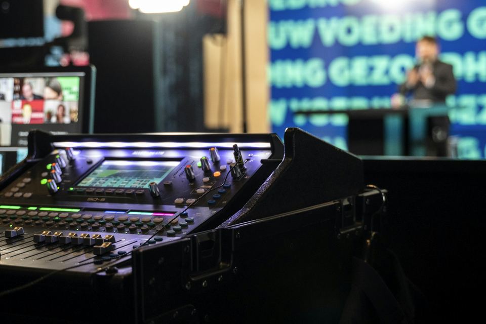 Digital audio console at front of house while a presenter speaks on stage